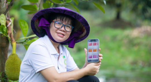 Nguyen Thi Kim Thoa sitting in an orchard showing a cellphone to the camera. She is wearing a large rimmed hat.