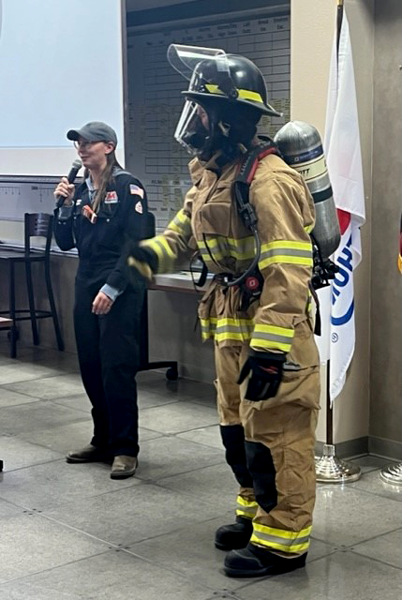 A person dressed in full fire-fighting PPE and a person next to them with a microphone.
