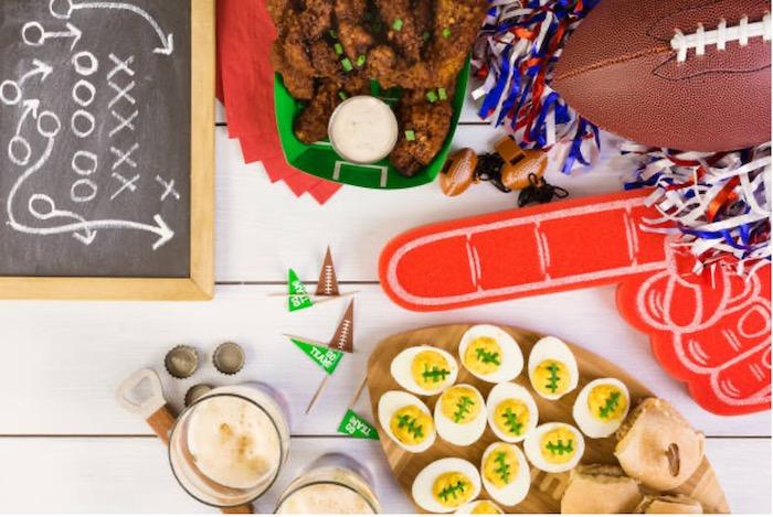 Party materials on a table for Super Bowl Sunday: Football, hard boiled eggs decorated like footballs, wings and a small chalk board with a play written in chalk.