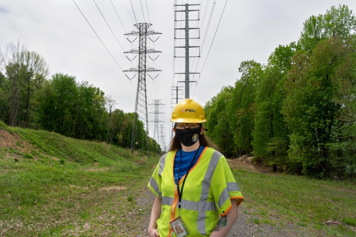 PSEG female employee standing under high tension power lines wearing protective gear, helmet and mask.