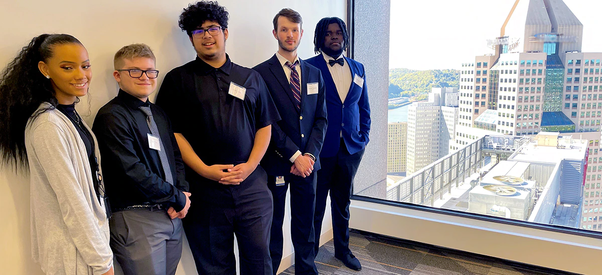 Five students in business style clothing standing in a hallway with a large window at the end overlooking a city landscape.