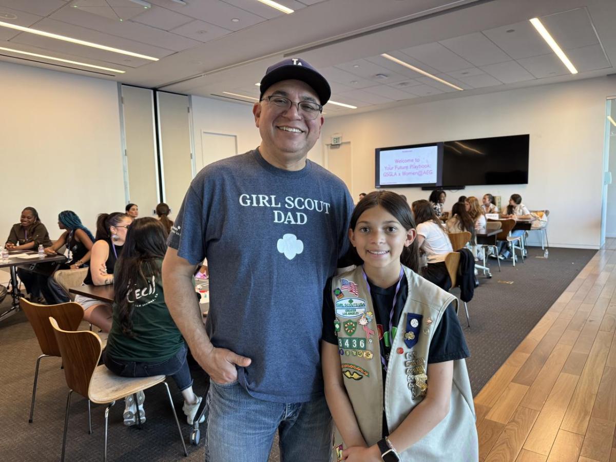 An adult and a youth participant stand together in a workshop space, with other attendees seated at tables behind them during a group program.