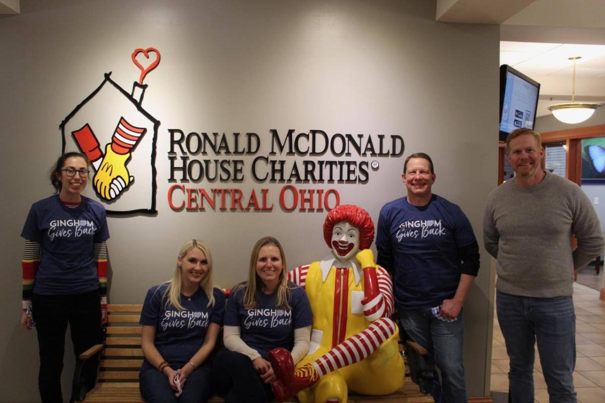 Five Bath & Body Works associates pose for a picture in front of a wall with a large logo of the Ronald McDonald House Charities of Central Ohio on it. Three associates are standing and two are sitting on a bench that includes a statue of Ronald McDonald with his leg crossed and showing a big smile.