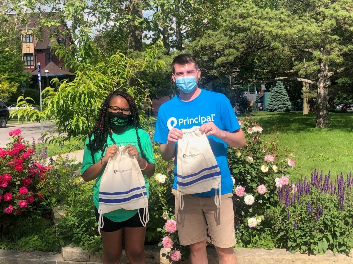 A young man and young woman; wearing face masks are holding a white bag in a garden.