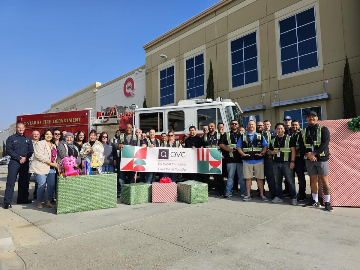 Qurate Retail Group team member's participating in a Together For Good volunteer event. A mix of team members from Qurate Retail Group are standing outside a QVC facility in front of a firetruck along with fire fighters. A few people are holding up a banner which reads "QVC Do What You Love, Love What You Do". They are also surrounded by wrapped gift boxes and holding children's toys as part of a holiday toy drive event.
