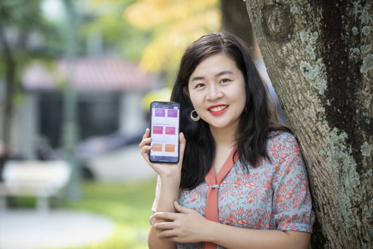 Quynh holding up a phone, leaning on a tree.