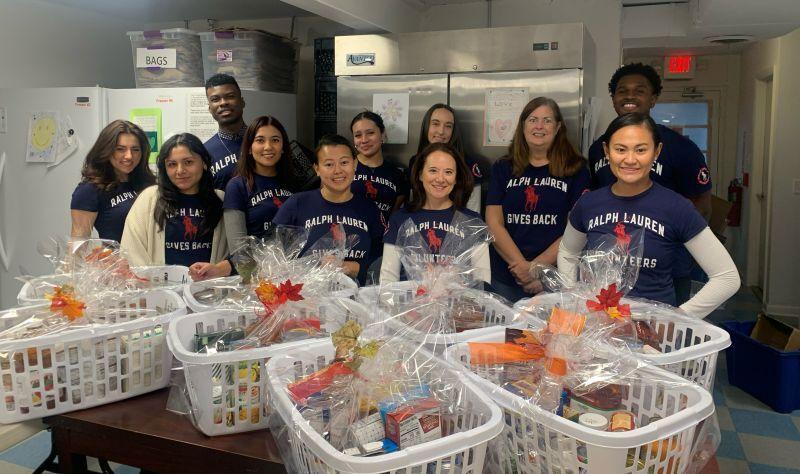 A group of people standing in a kitchen, large baskets of food items in front of them. All wearing "Ralph Lauren Volunteer" t-shirts.