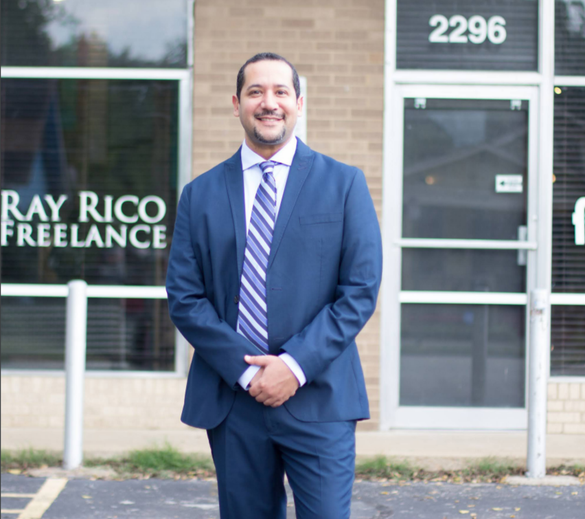 Ray Rico standing outside their business. Wearing a blue suit and striped tie.