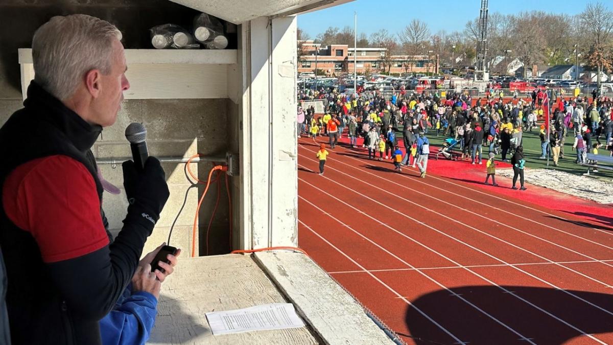 Robert McNamee in a booth with a microphone, looking out to a race track filled with participants.