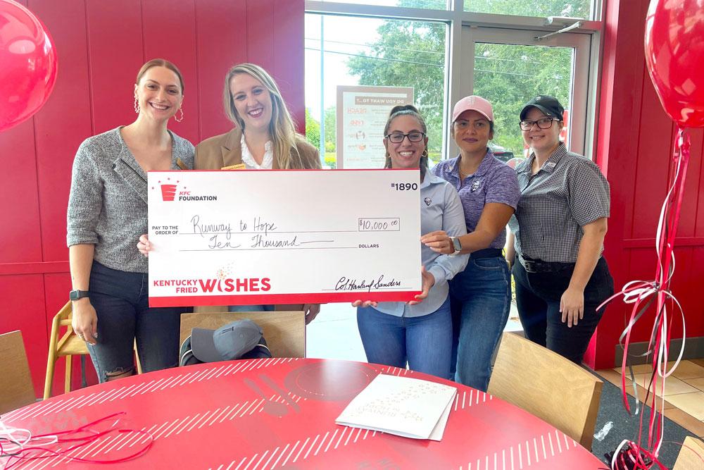 Group of people stood in front of a red wall holding a Kentucky Fried Wishes funding cheque