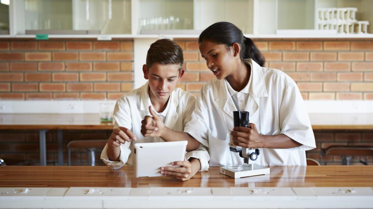 Two young students in a lab looking at a tablet device while another holds a microscope