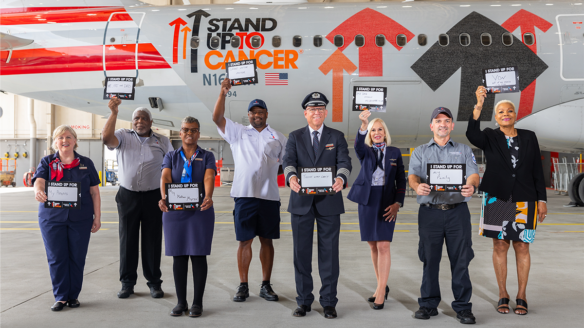 A row of employees holding up signs in front of a plane with "Stand up to Cancer" decals.