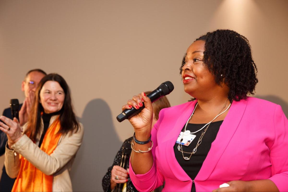 Salah Goss with a microphone, others to her side clapping and smiling.