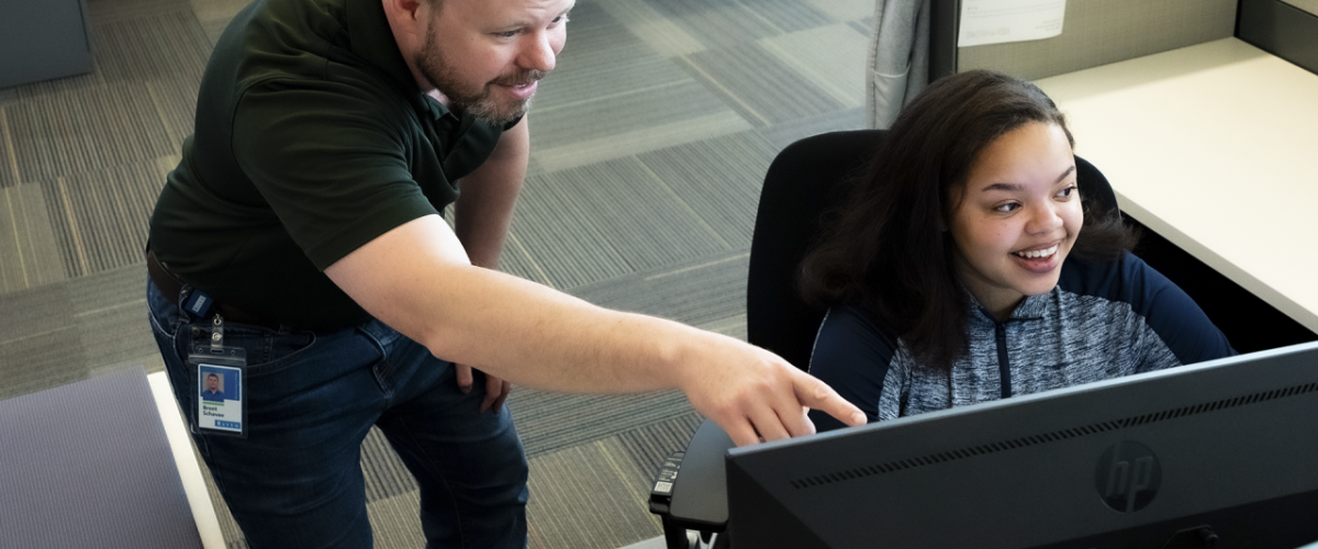 The Women Coding Success for Raven Person sat at a desk in front of a computer screen with someone stood next to them pointing at the monitor