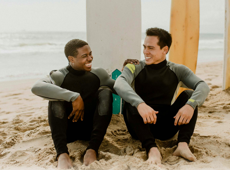 two men sitting on the beach wearing wet suites for surfing