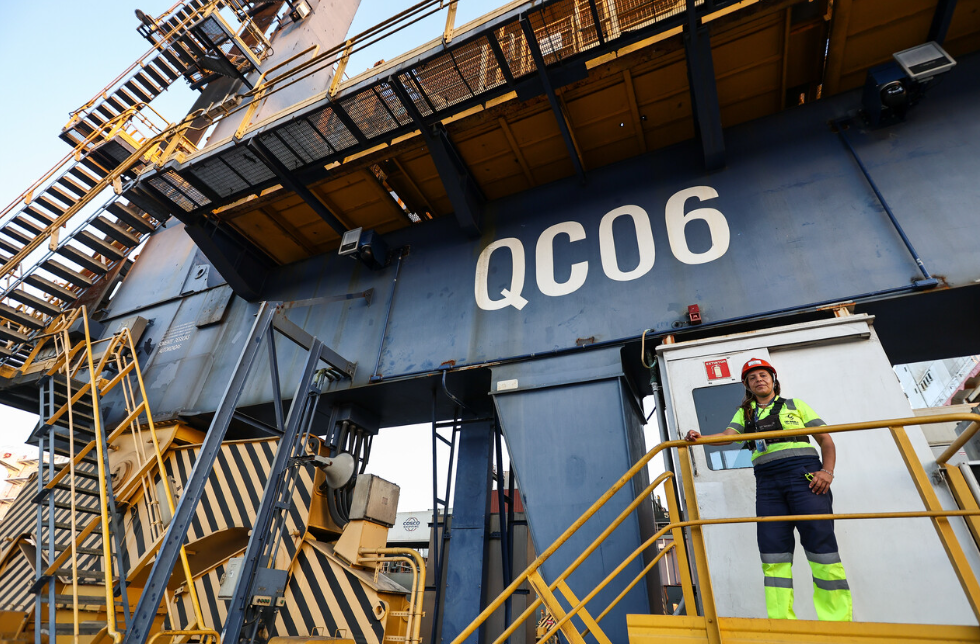 Female port worker wearing a hard hat stands on a platform beside a large quay crane labeled QC06 at DP World’s Santos terminal in Brazil.