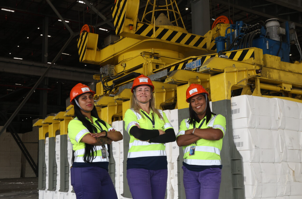 Three women wearing high-visibility uniforms and hard hats stand in front of large cargo-handling machinery inside a DP World warehouse facility in Brazil.