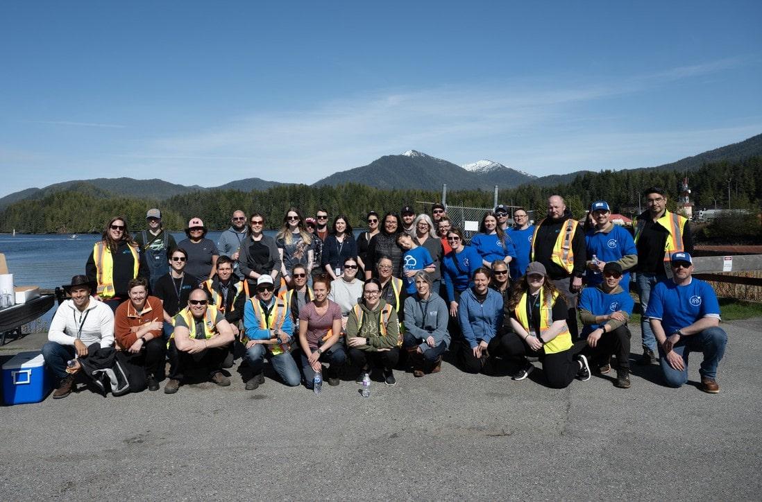 A large group of DP World employees and community volunteers posing together near the coastline with mountains and water in the background during a cleanup event.