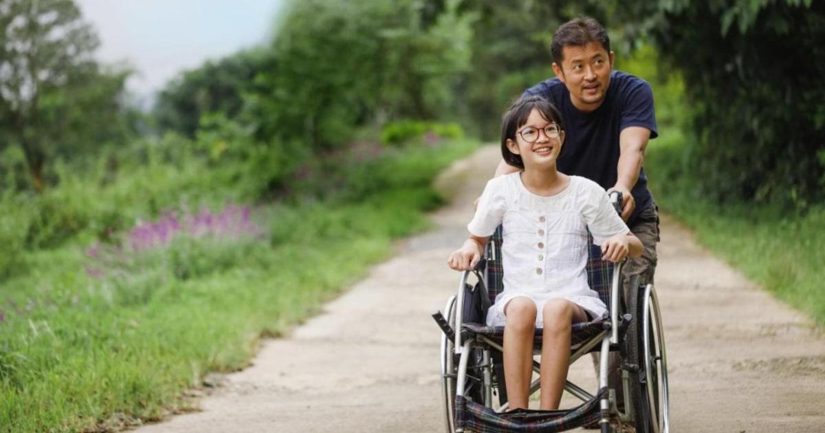 An adult pushing a child in a wheelchair on a sandy path with lush greenery on the sides.