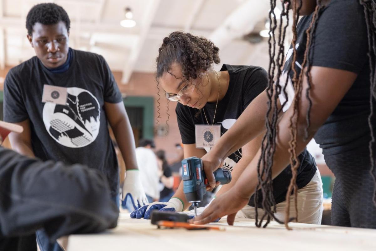 Person using a tool with another person helping and someone watching at the side