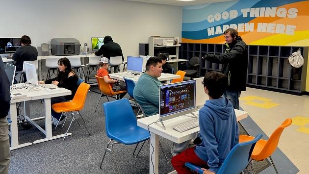 A group of children sitting at desks with computers. A row of cabinets for coats and bags on the side wall under a mural "Good Things Happen Here."