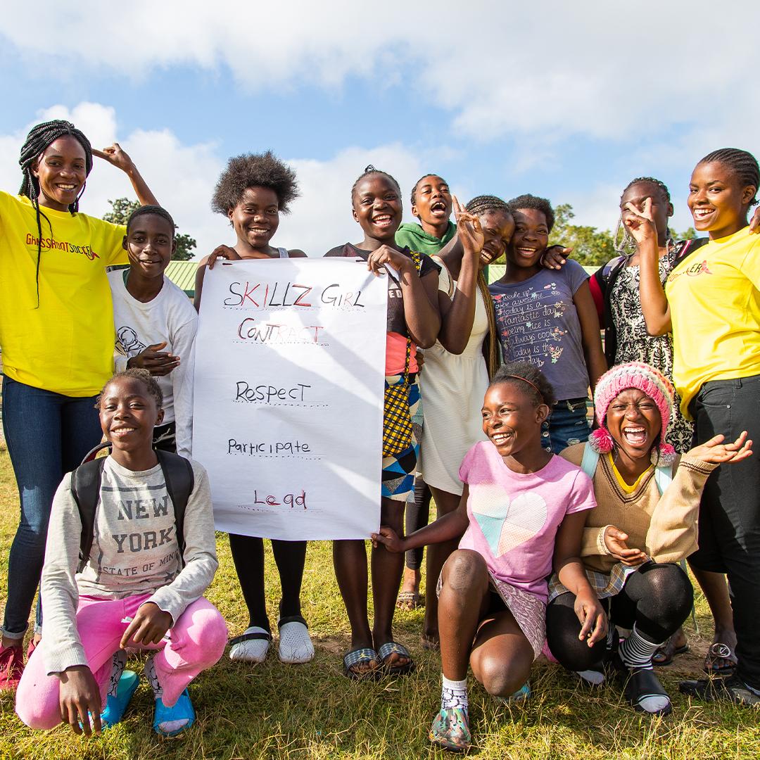 A group of youths smiling, posing for the camera on an open field, holding a sign "Skills Girl contract."