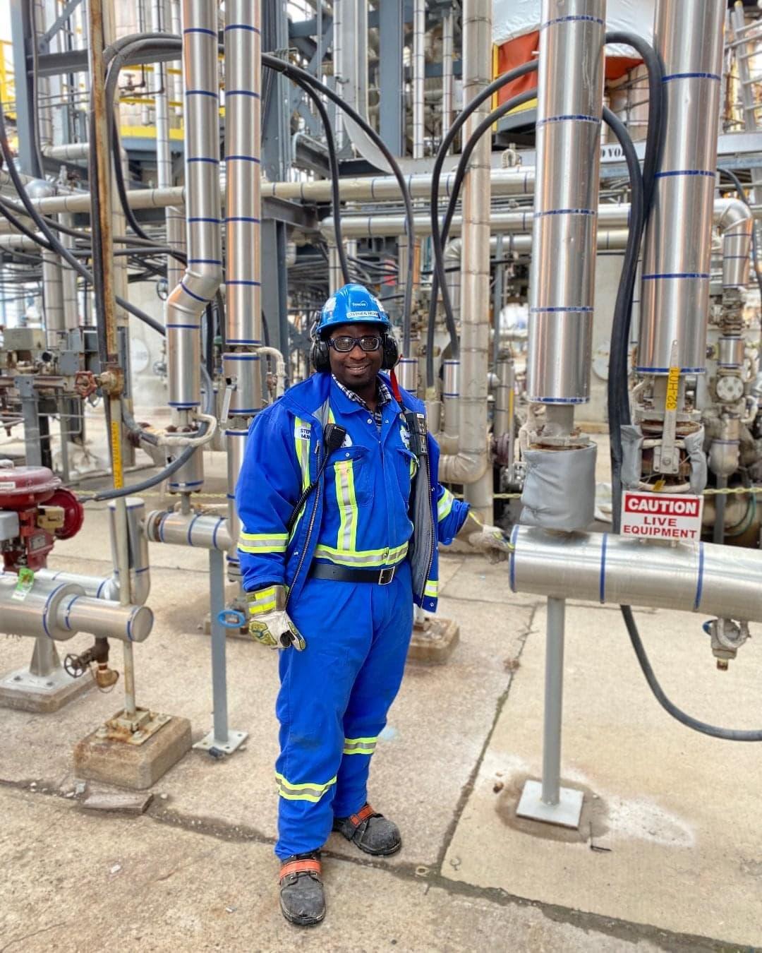 Stephen Muinda standing in an industrial complex, wearing protective clothing, hard hat, glasses, and ear muffs.