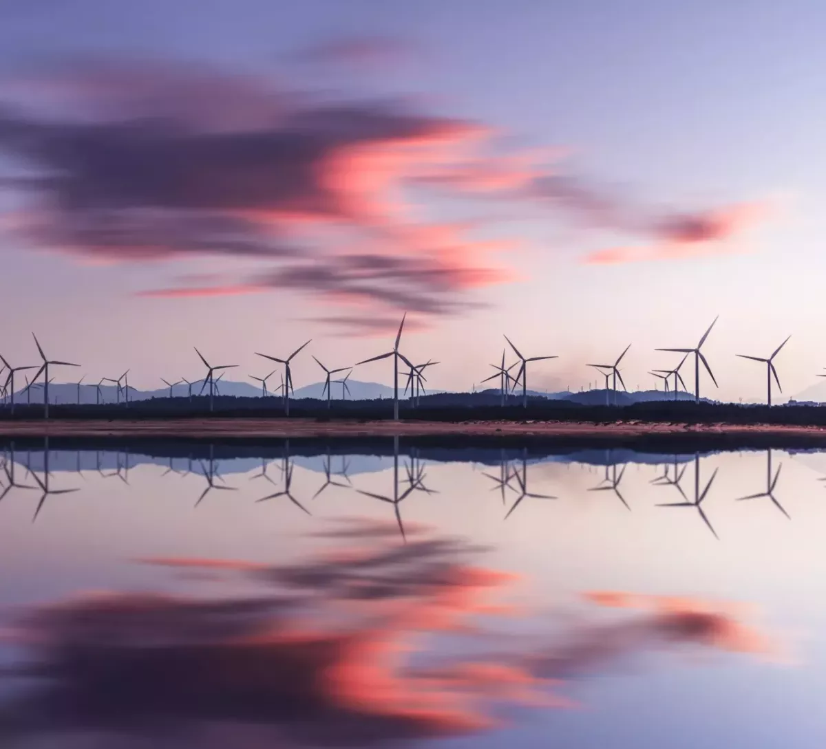 Rows of wind turbines in the distance, an open body of water in front reflecting clouds in a sunset.