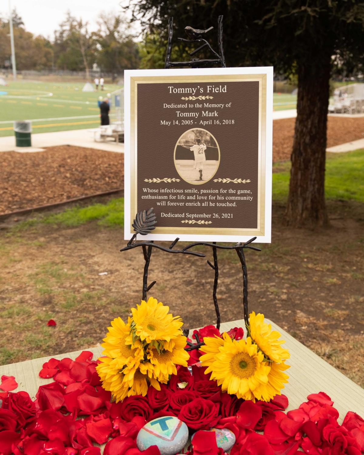 A commemorative plaque for Tommy Mark is positioned by "Tommy's Field" surrounded by flowers. 
