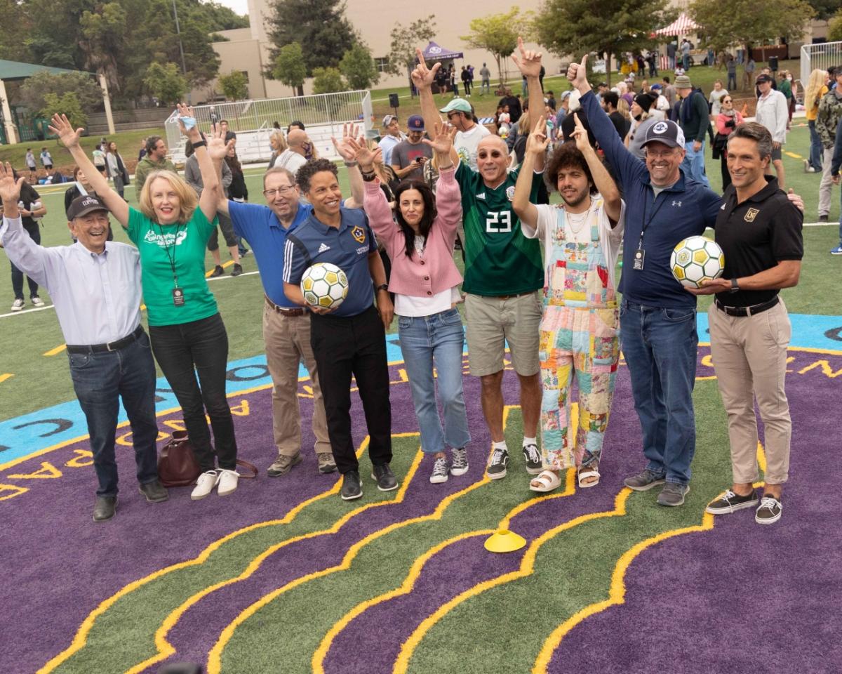 Members of the Mark Family, community members and representatives of the LA Galaxy and LAFC celebrate with a cheer and their hands in the air in the middle of "Tommy's Field." 