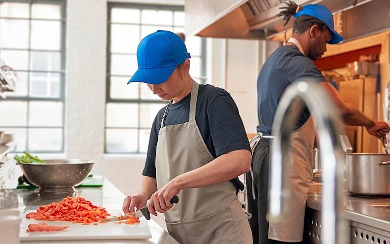 Two workers are shown preparing food in a restaurant. Both are wearing blue caps and aprons.