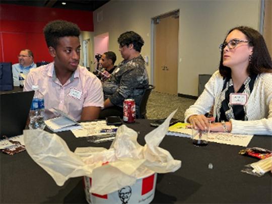 Truman and another talking at a table in a room with others. KFC bucket central.