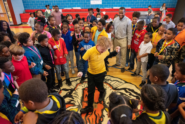 a large group of kids in a gymnasium, circled around one who is dancing in the middle.