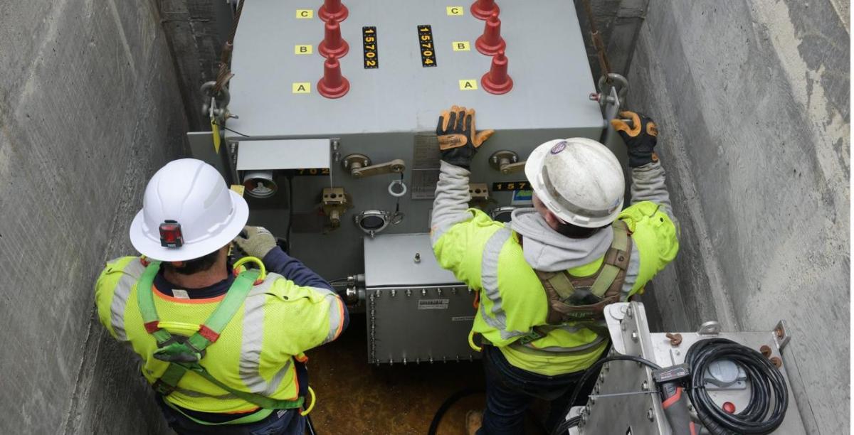 Two people in protective uniform working on a large electrical unit underground.