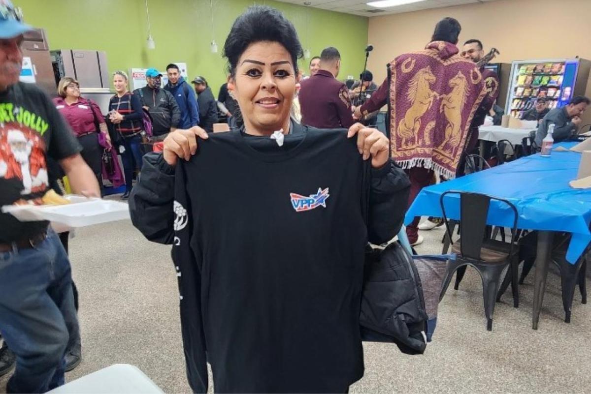 a person holding up a black long-sleeve shirt with VPP on the front pocket. Others in the room walking around.