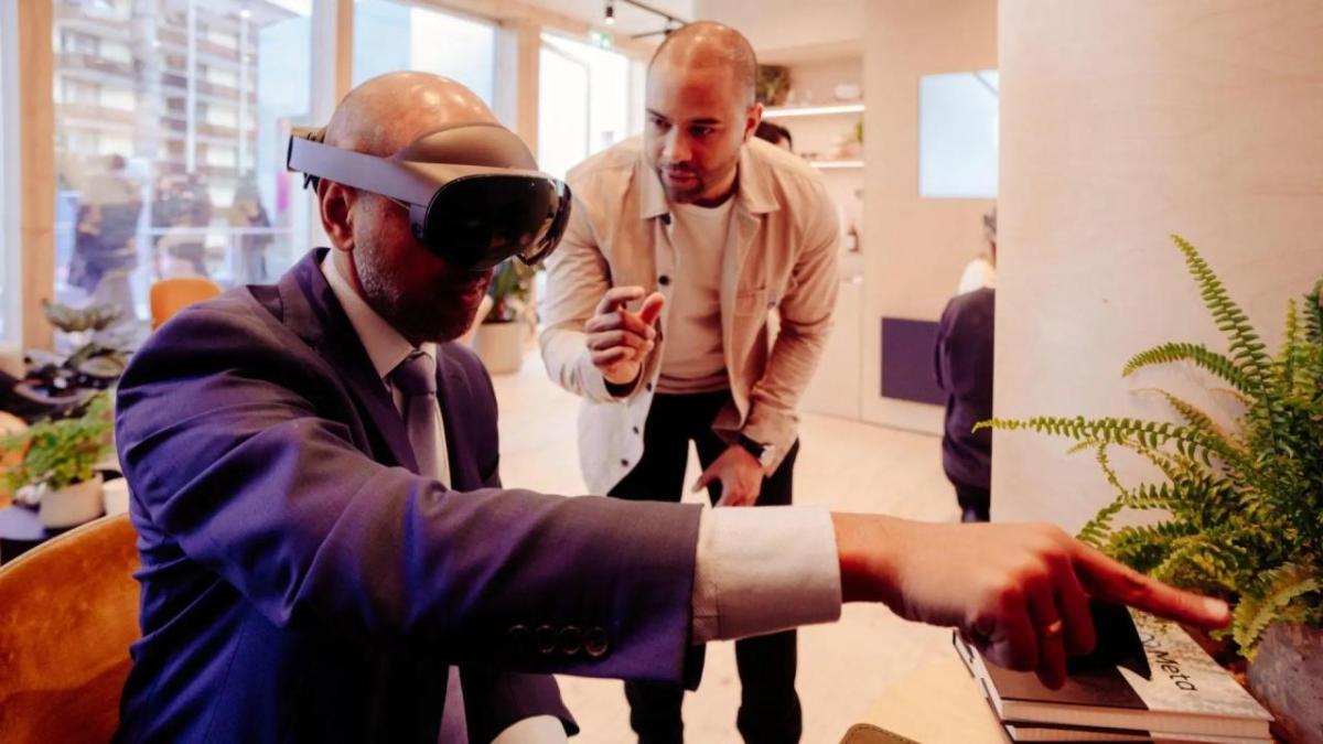 A seated person in a suit using a VR headset, pointing off screen. A person standing next to them talking.