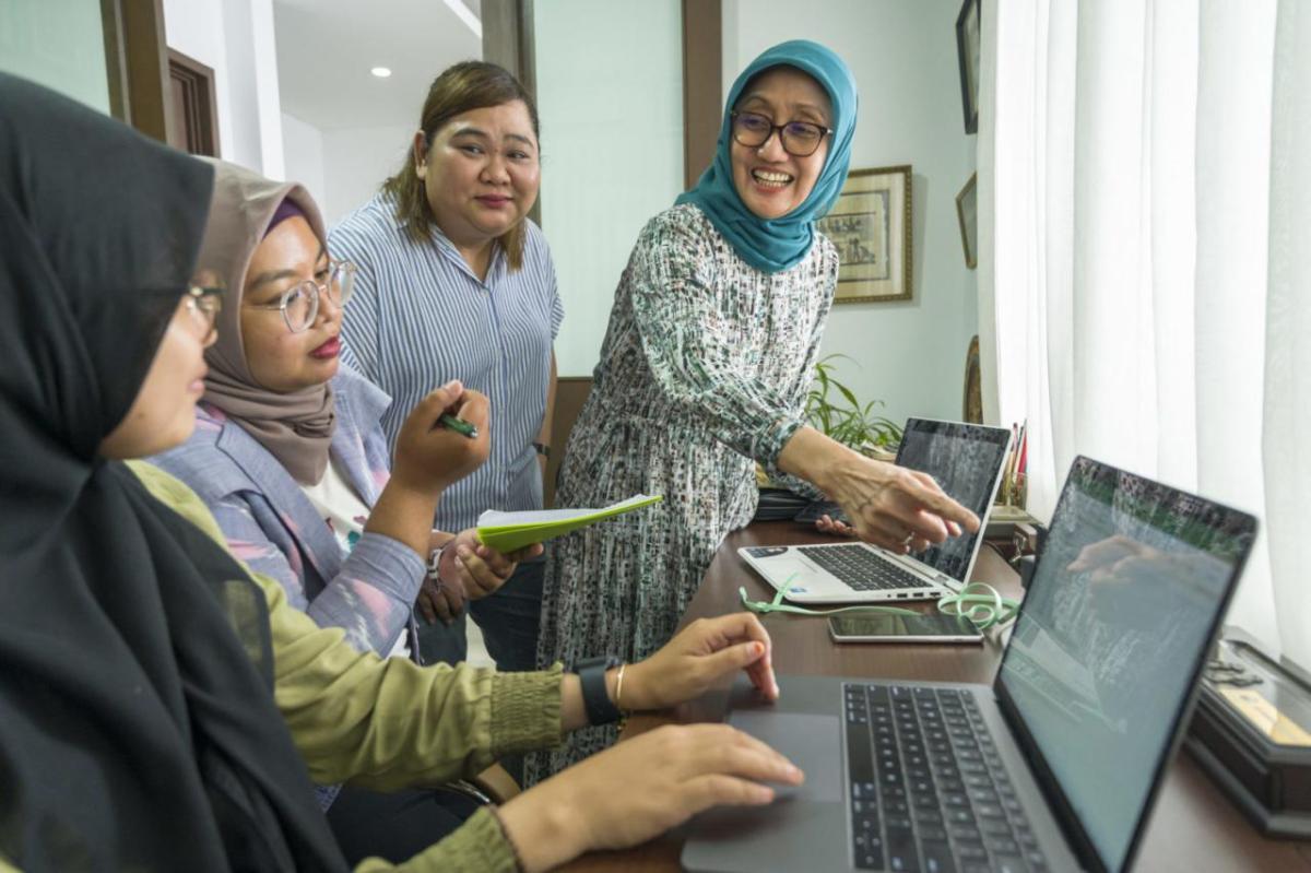 Four women talking as one uses a laptop at a desk.