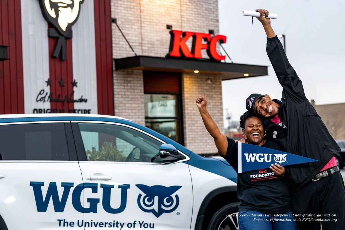 Two people smiling with one arm raised. One holds a WGU pendant. Standing in front of a car with WGU and logo on the side and all in front of a KFC restaurant.