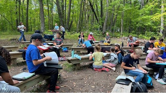 a group of students on benches outdoors in a wooded area, looking at open books.