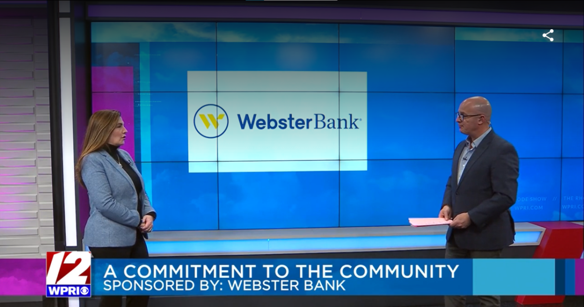 Two people standing in front of a large digital screen, one with papers, the other being interviewed.