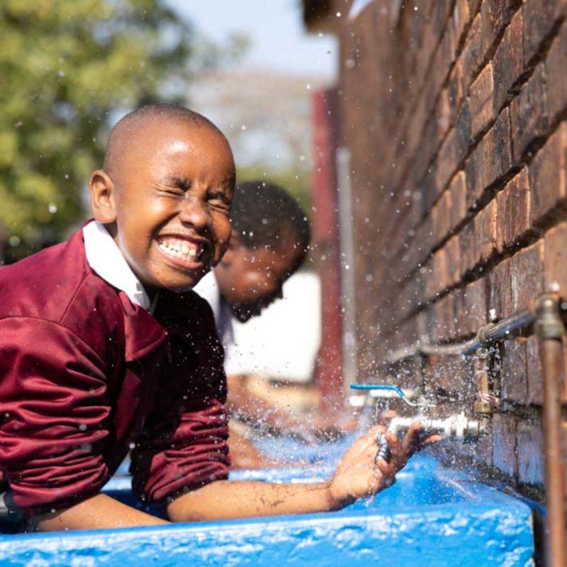 A smiling child splashing in an outdoor faucet.