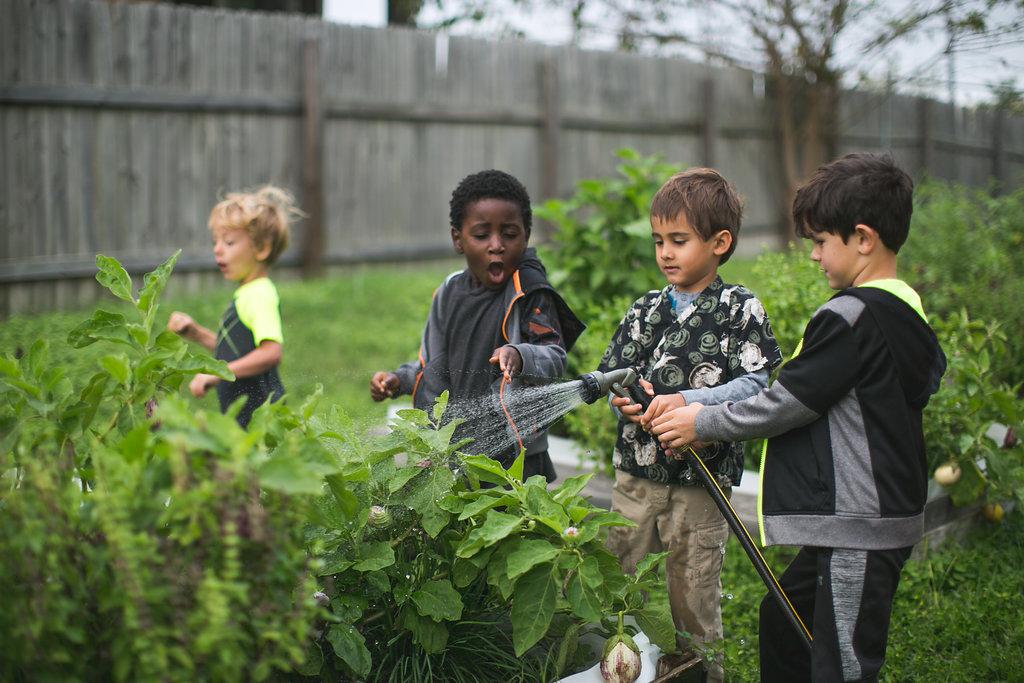 Children in a garden, one using a hose to water plants.