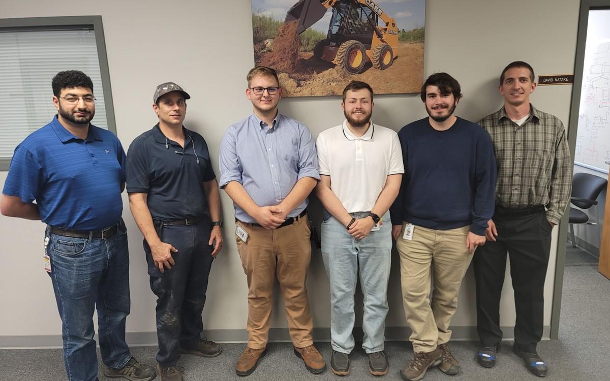 Six people posed in a hallway in front of an image of a case front-loader.