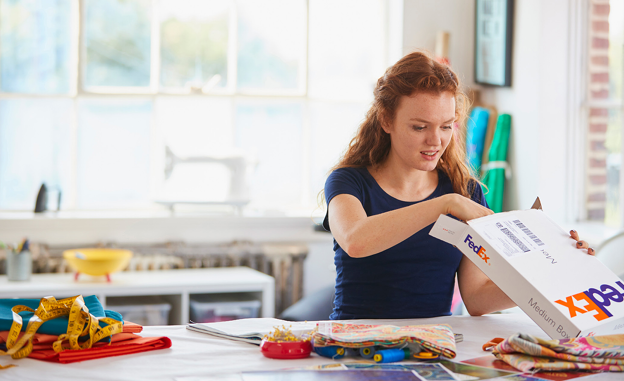 a person in a bright room, sitting at a large table with craft items, opening a FedEx box