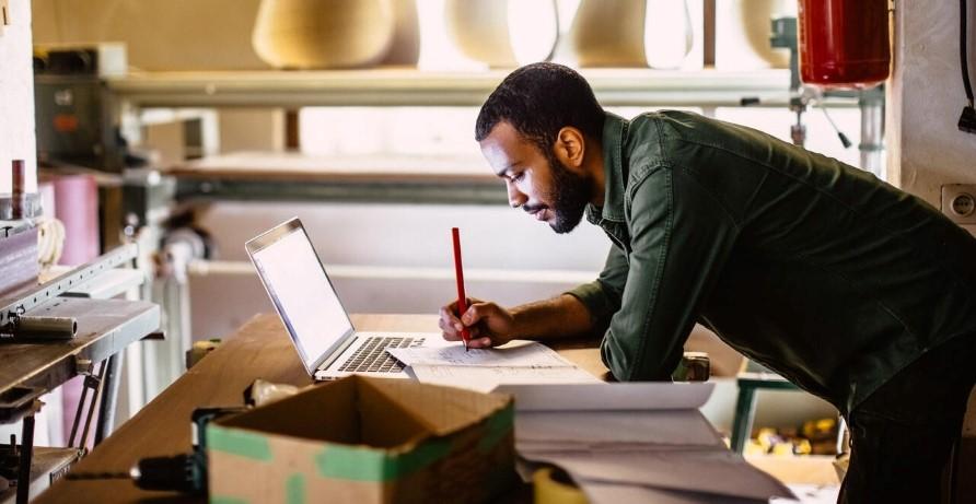 A person leant on a desk writing in a book that it resting on a laptop