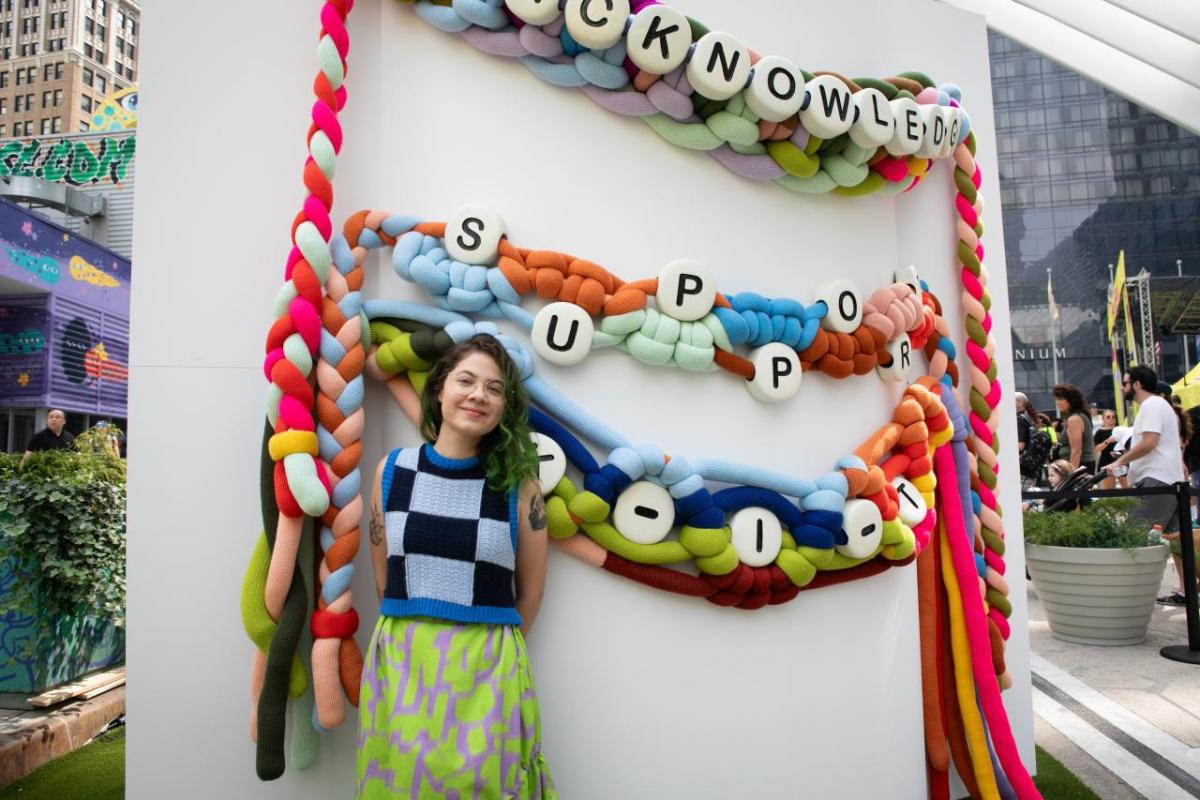 woman standing in front of giant friendship bracelets with the words "acknowledge" and "support"