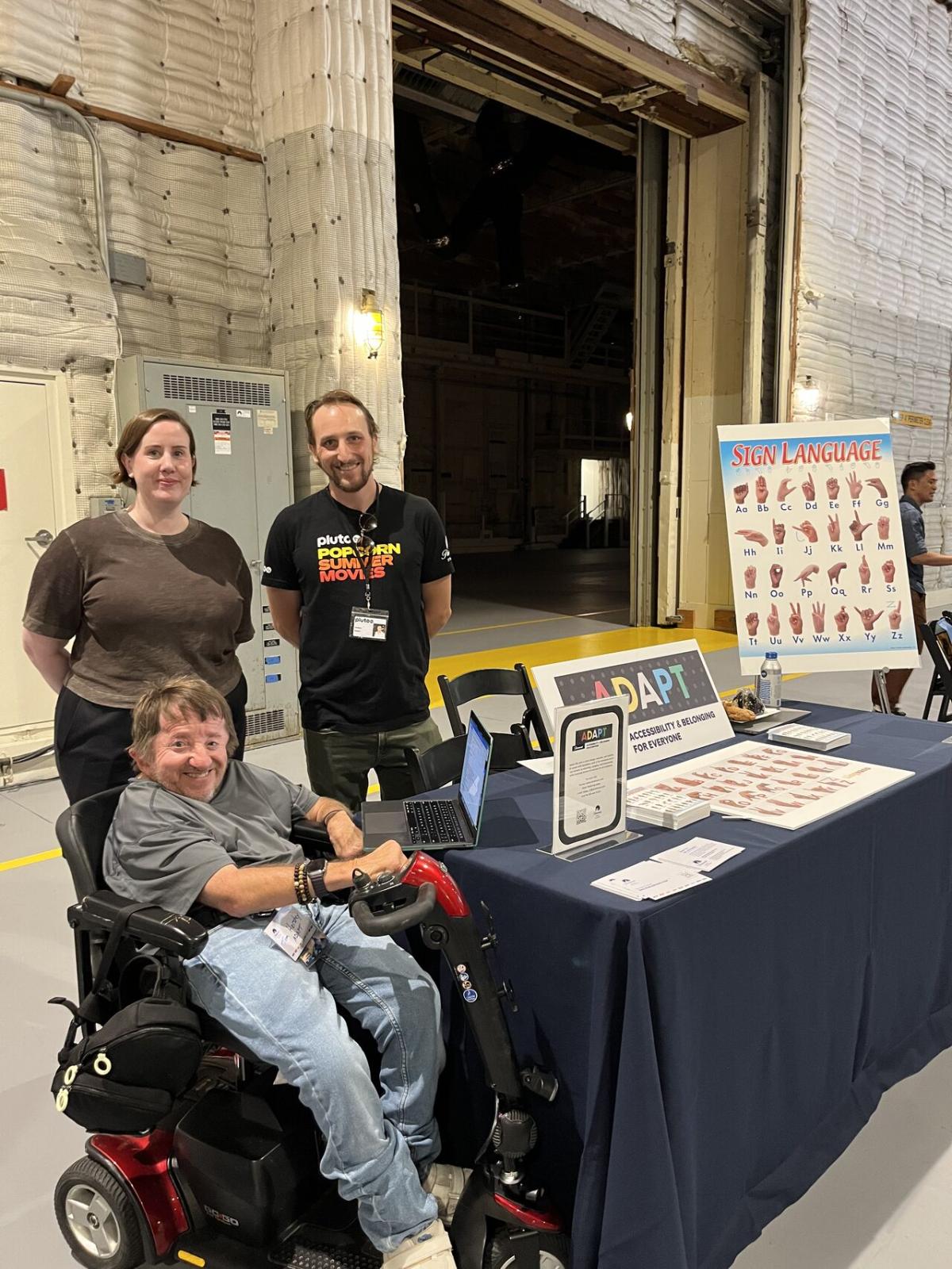 Three people by a table with ADAPT literature and an asl board.