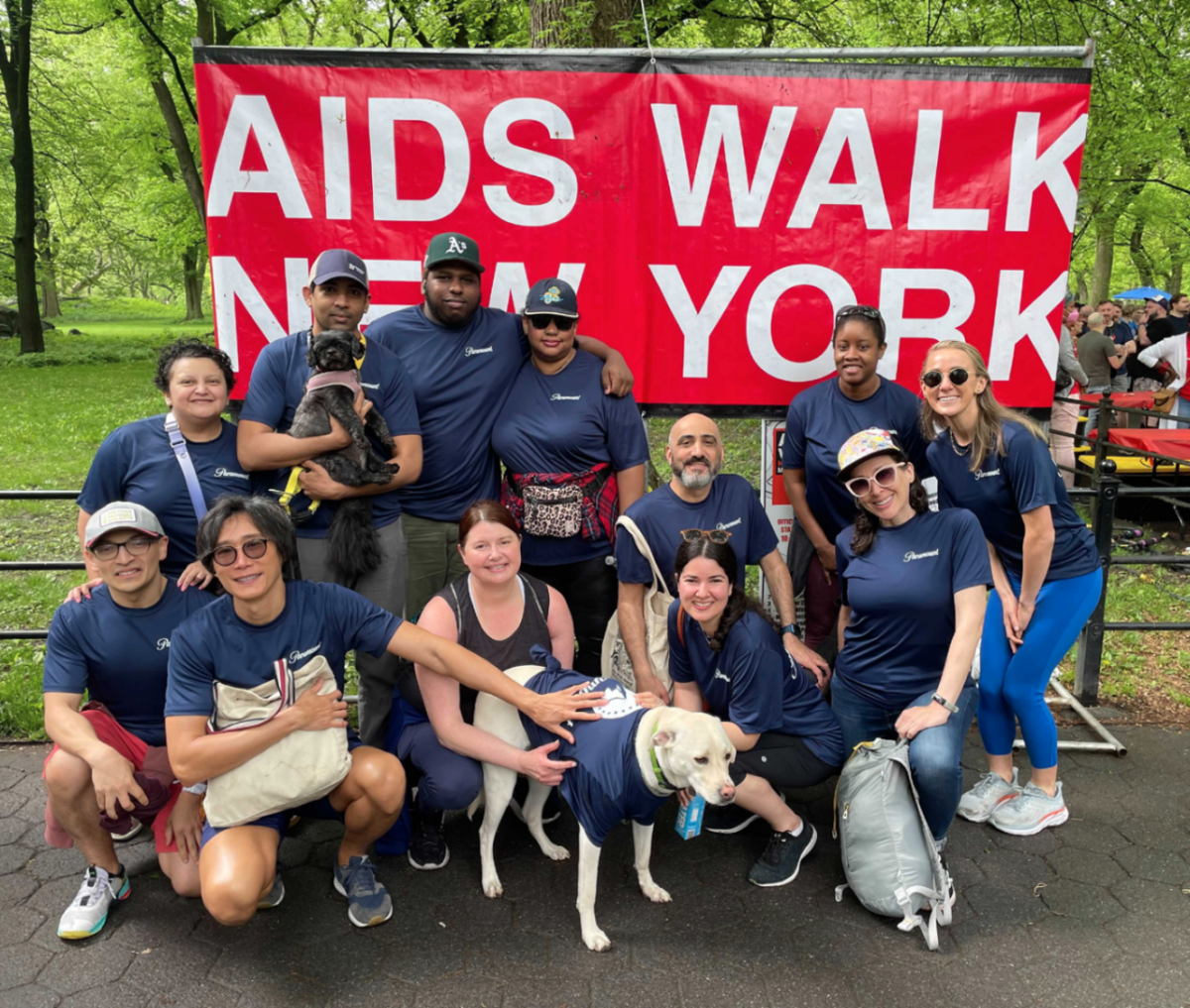 AIDS Walk 2022 A group of people, some sitting in front of others standing. Two dogs in front getting pet. All wearing matching t-shirts (even the good dog). "AIDS WALK NEW YORK" on a banner behind them.
