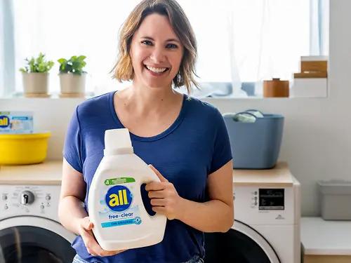 A smiling person holding a bottle of all detergent in front of a washer and dryer.
