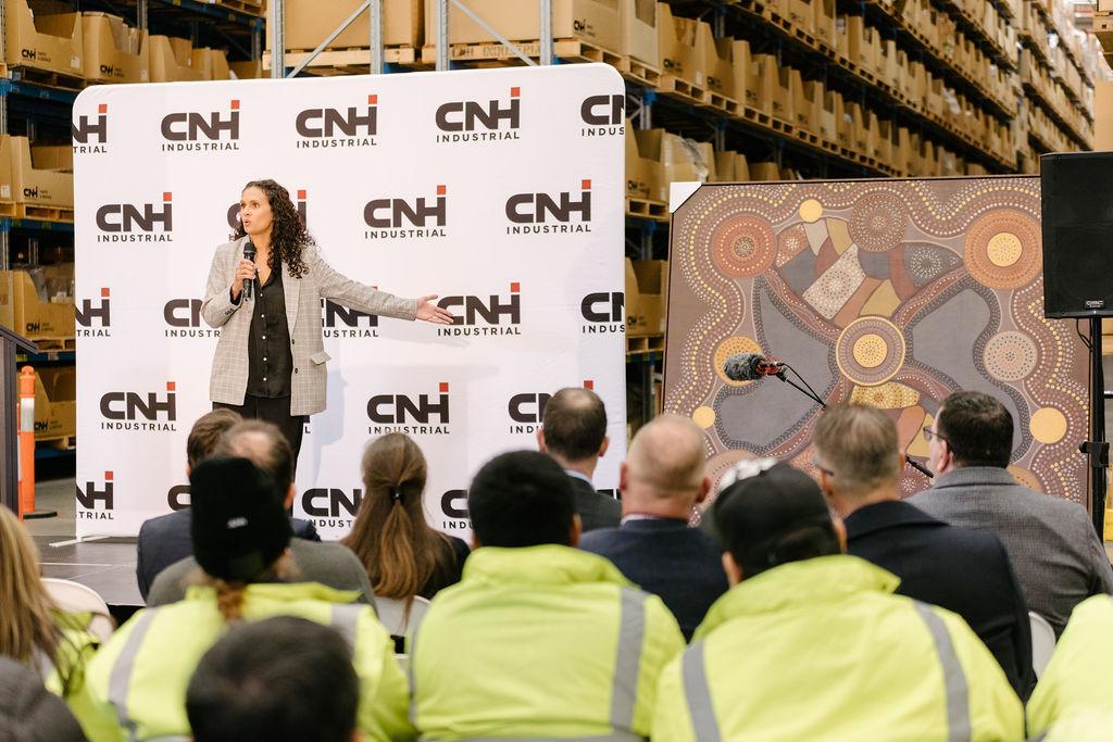 A person presenting next to artwork in front of a crowd, some in safety clothing. all in a warehouse.